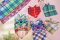 A styled flat-lay of multiple colorful plaid bandana neckties arranged on a pink and beige surface, with a matching plaid fabric strip in the upper left corner.