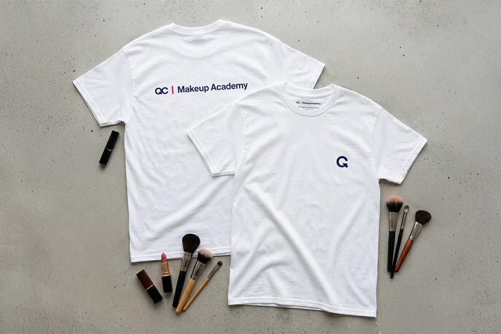 Two white t-shirts with makeup academy branding on a gray surface with makeup brushes.