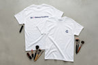 Two white t-shirts with makeup academy branding on a gray surface with makeup brushes.