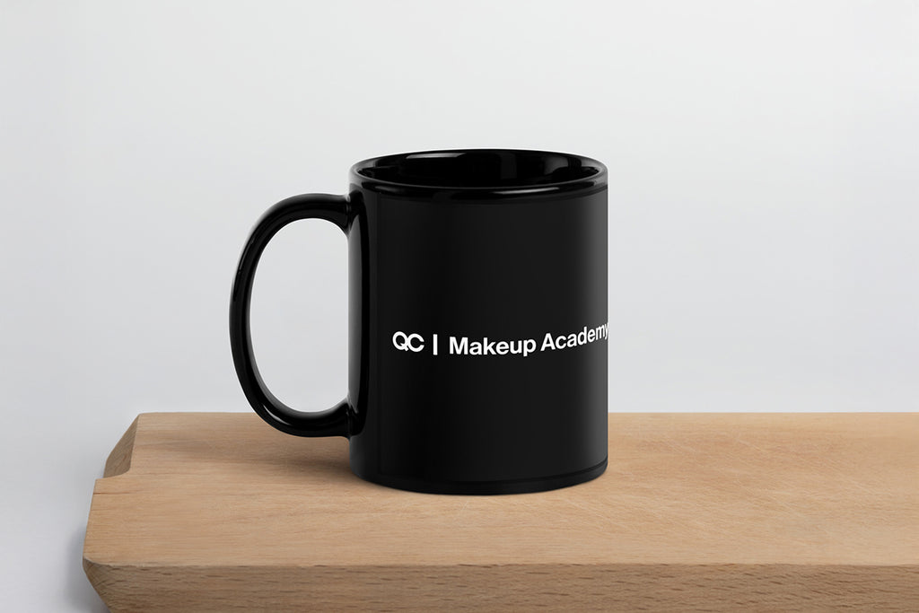Black mug with 'QC Makeup Academy' text on a wooden surface and light gray background
