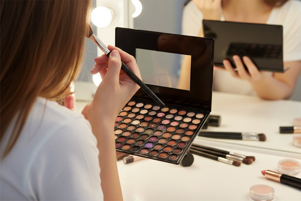 Person applying makeup with a palette and brushes in a well-lit room.