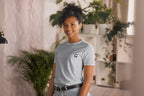 Person wearing a gray t-shirt with a QC logo in an indoor setting with plants