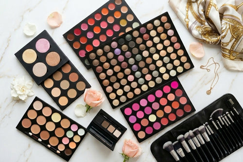 Collection of makeup palettes and brushes on a marble surface with flowers and jewelry.