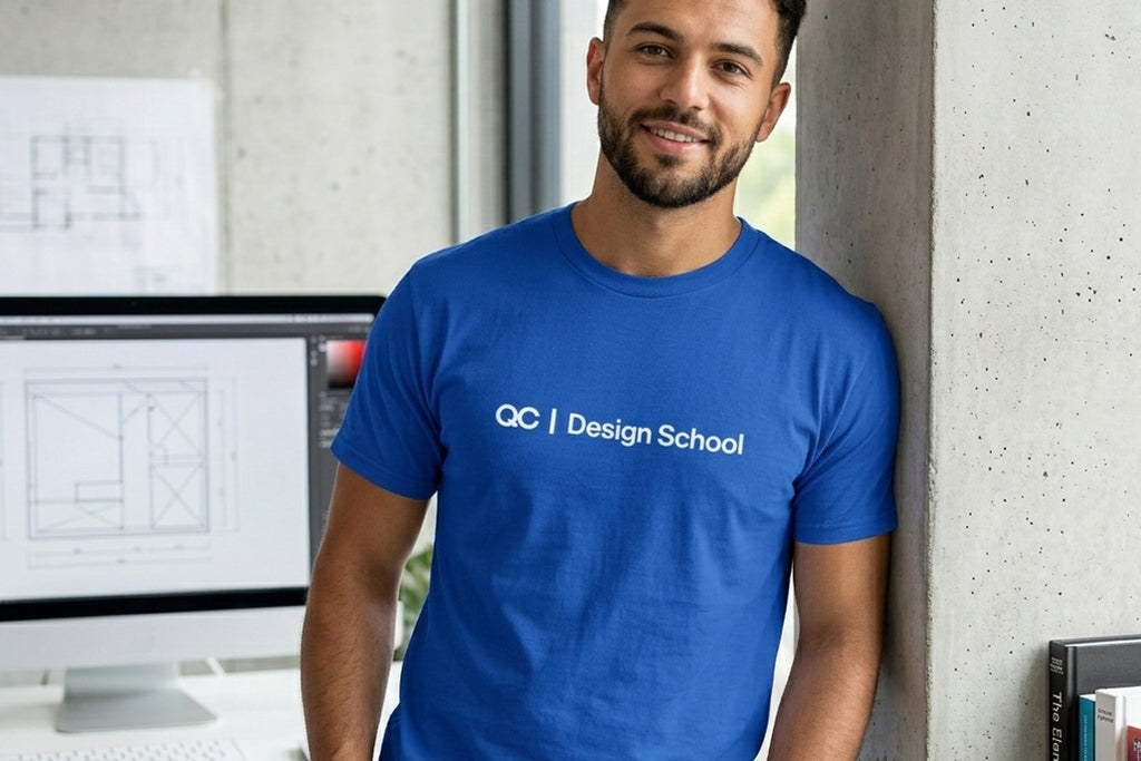 A medium shot of a smiling man with a beard, leaning casually against a textured concrete wall in a modern architectural design studio. He is wearing a royal blue t-shirt with 'QC | Design School' printed in white text across the chest. The background shows a computer screen displaying blueprints, books, and architectural drawings.