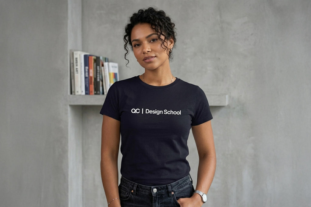 Medium shot of a woman with curly dark hair standing against a raw, textured concrete wall. She is wearing a black crew-neck t-shirt with the text "QC | Design School" in white across the chest. A row of colorful books is on a shelf behind her. She has a hand in her pocket and is wearing a silver watch and necklace.