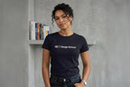 Medium shot of a woman with curly dark hair standing against a raw, textured concrete wall. She is wearing a black crew-neck t-shirt with the text "QC | Design School" in white across the chest. A row of colorful books is on a shelf behind her. She has a hand in her pocket and is wearing a silver watch and necklace.