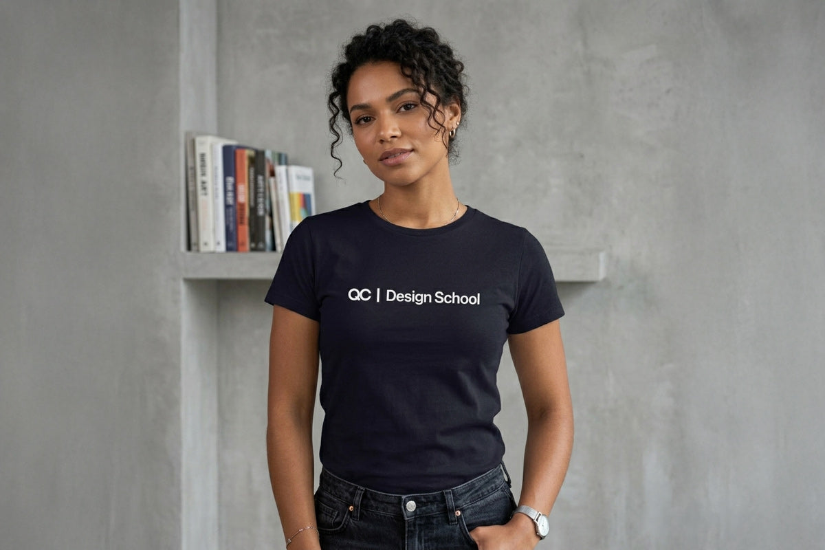 Medium shot of a woman with curly dark hair standing against a raw, textured concrete wall. She is wearing a black crew-neck t-shirt with the text "QC | Design School" in white across the chest. A row of colorful books is on a shelf behind her. She has a hand in her pocket and is wearing a silver watch and necklace.