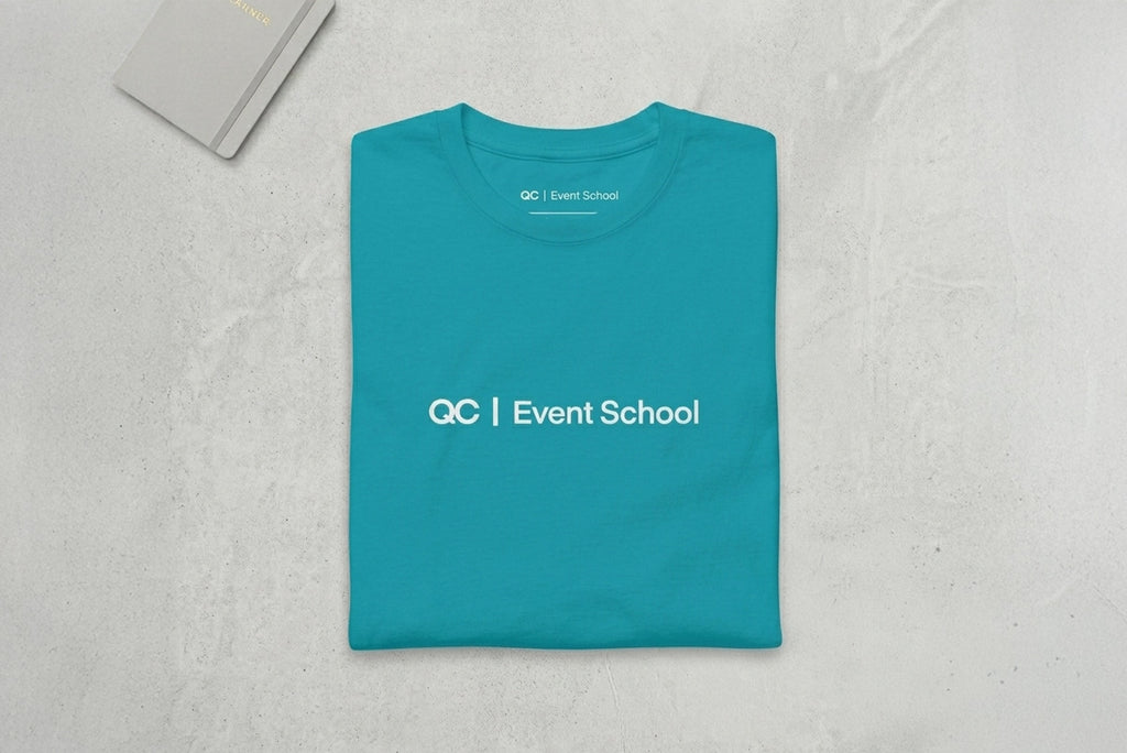 Teal t-shirt with 'QC Event School' text on a light gray textured surface