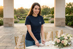 An image of a smiling young woman with long brown hair, looking directly at the camera while standing at and setting a formal outdoor dining table. She wears a navy blue t-shirt with a white 'G' logo and blue jeans. A rose tattoo is visible on her left forearm. She is reaching down with one hand to place cutlery. The table is elegantly set with china, various stemware, silver, and a detailed floral centerpiece. Behind her are two gold Chiavari chairs with fabric sashes.