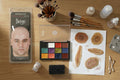 Makeup palette with color swatches, brushes, a sponge, and a bald cap on a wooden surface.