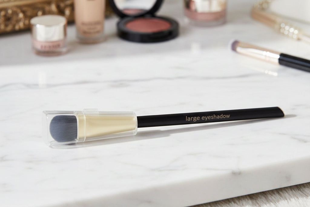 Makeup brush with black handle and gray bristle on a marble surface with cosmetic products in the background.