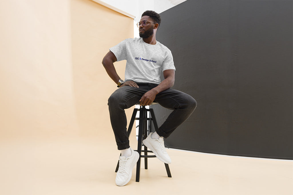 Man sitting on a stool wearing a gray t-shirt that reads "QC Design School", black pants, and white sneakers in a minimalistic setting.