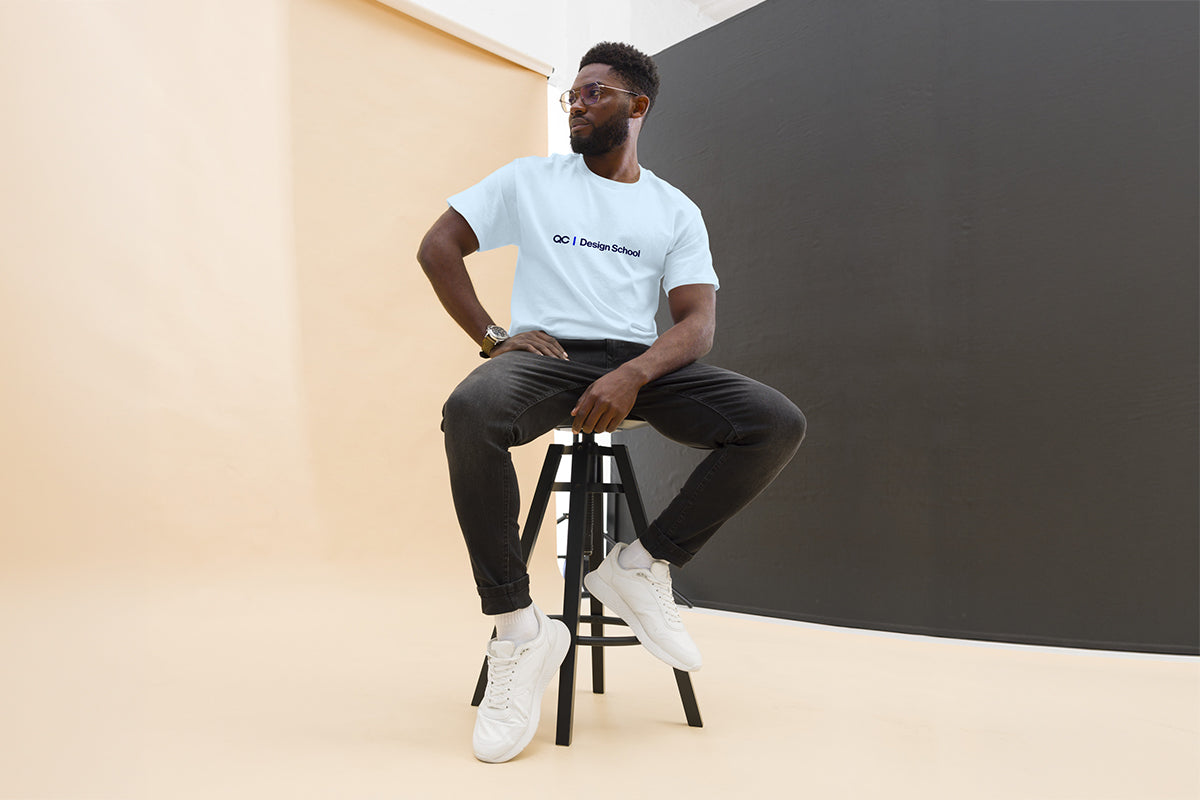 Man sitting on a stool wearing a light blue t-shirt that reads "QC Design School" and dark pants against a plain background