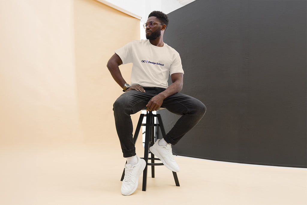 Man sitting on a stool wearing a beige t-shirt that reads "QC Design School" and black pants in a minimalistic setting.
