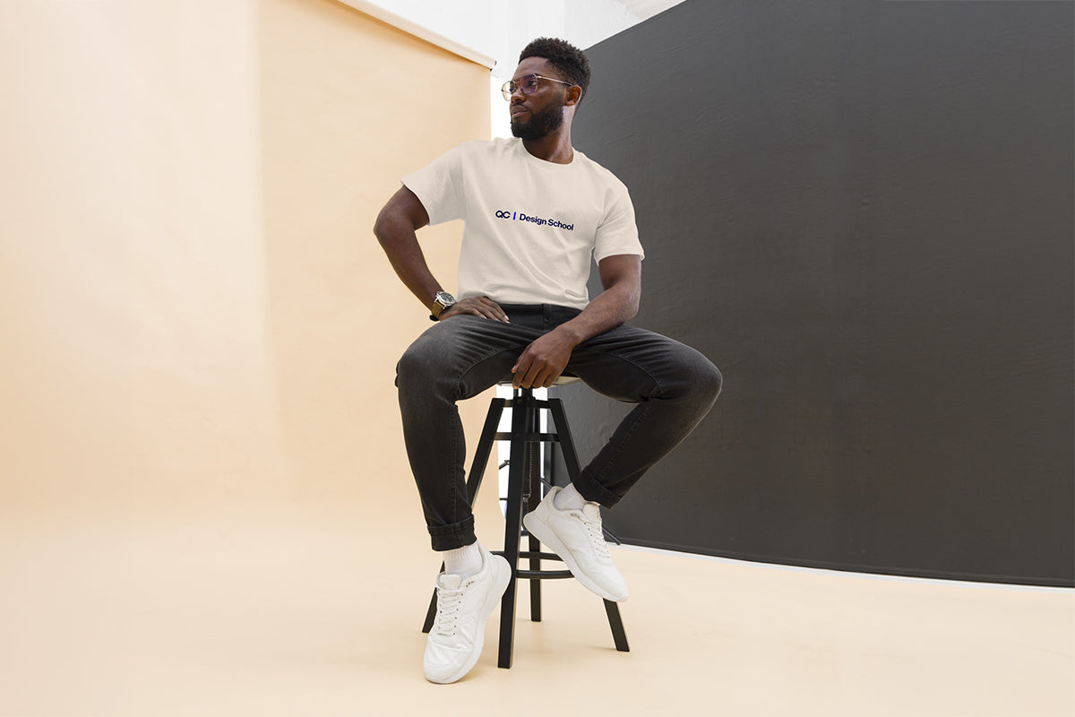 Man sitting on a stool wearing a beige t-shirt that reads "QC Design School" and black pants in a minimalistic setting.