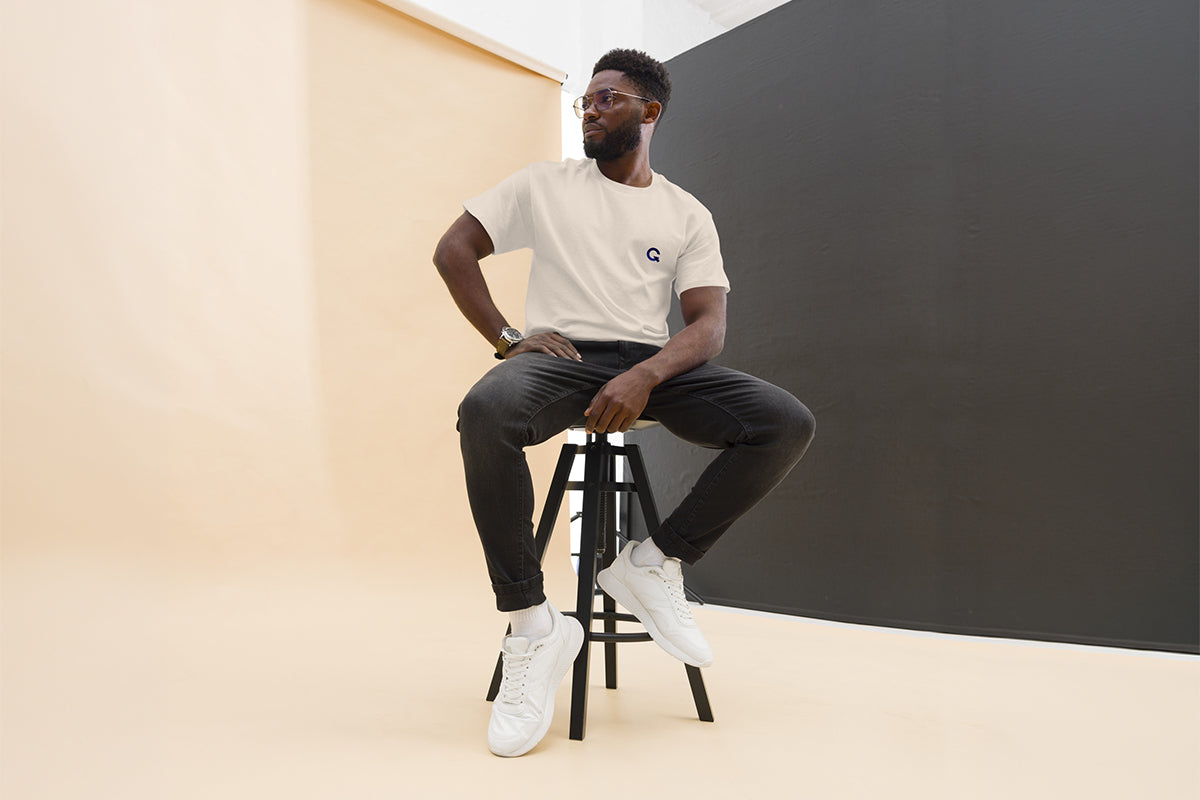 Man sitting on a stool wearing beige QC Design School shirt against a plain background