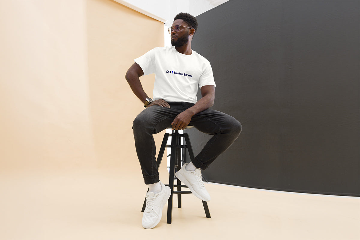 Man sitting on a stool wearing a white t-shirt that reads "QC Design School", black pants, and white sneakers against a plain background.