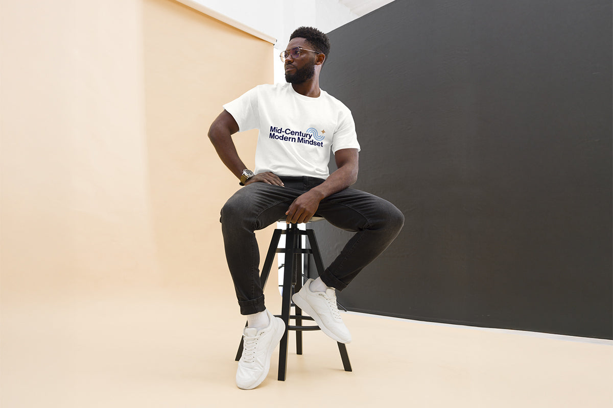 Person sitting on a stool wearing a white t-shirt that reads "Mid-Century Modern Mindset" and dark pants against a plain background