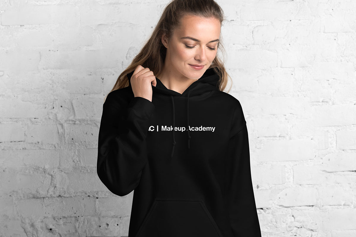 Woman wearing black hoodie that reads "QC Makeup Academy". She's standing against a white brick wall.