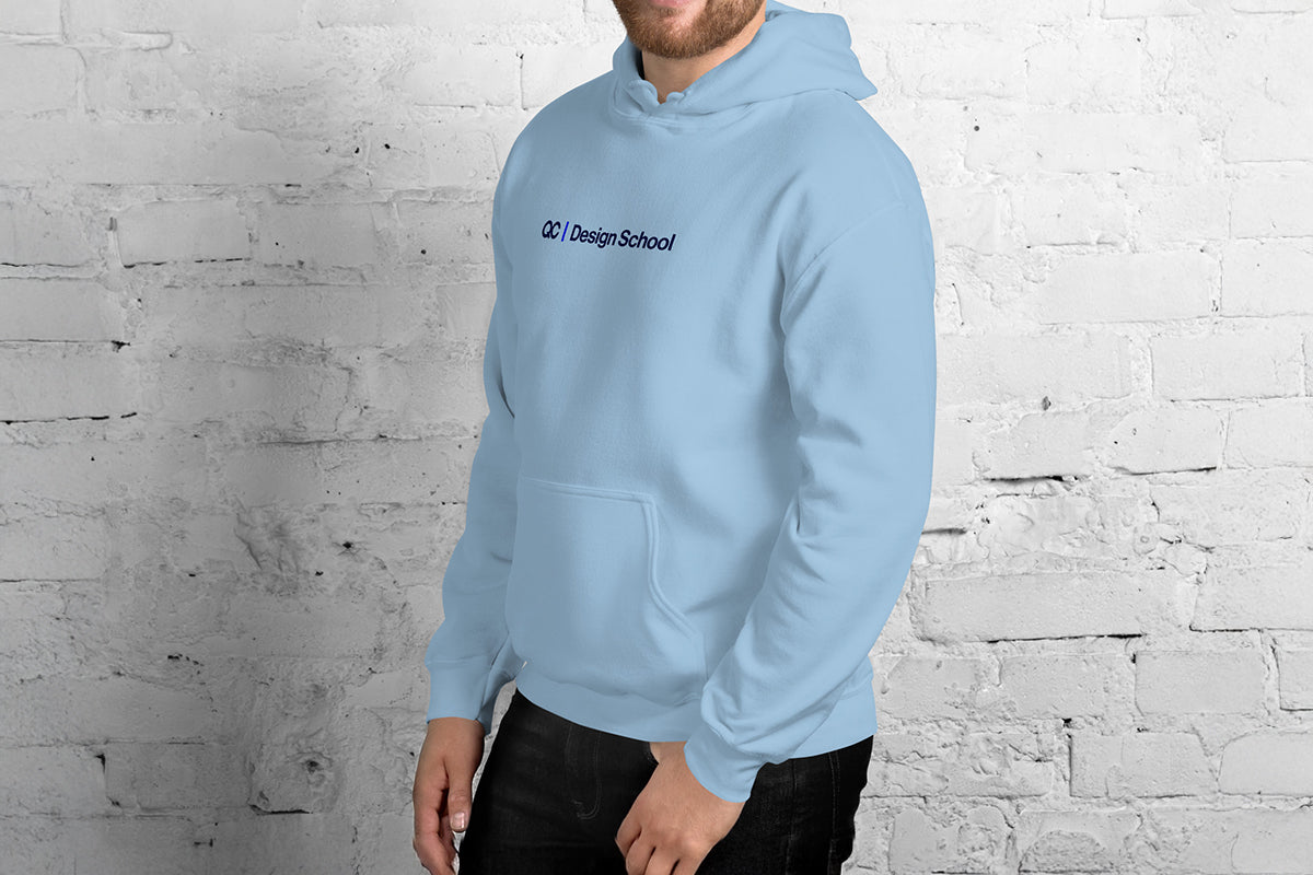 Person wearing a light blue hoodie with 'QC Design School' text against a white brick wall.