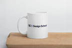 White mug with 'QC Design School' text on a wooden surface and light gray background