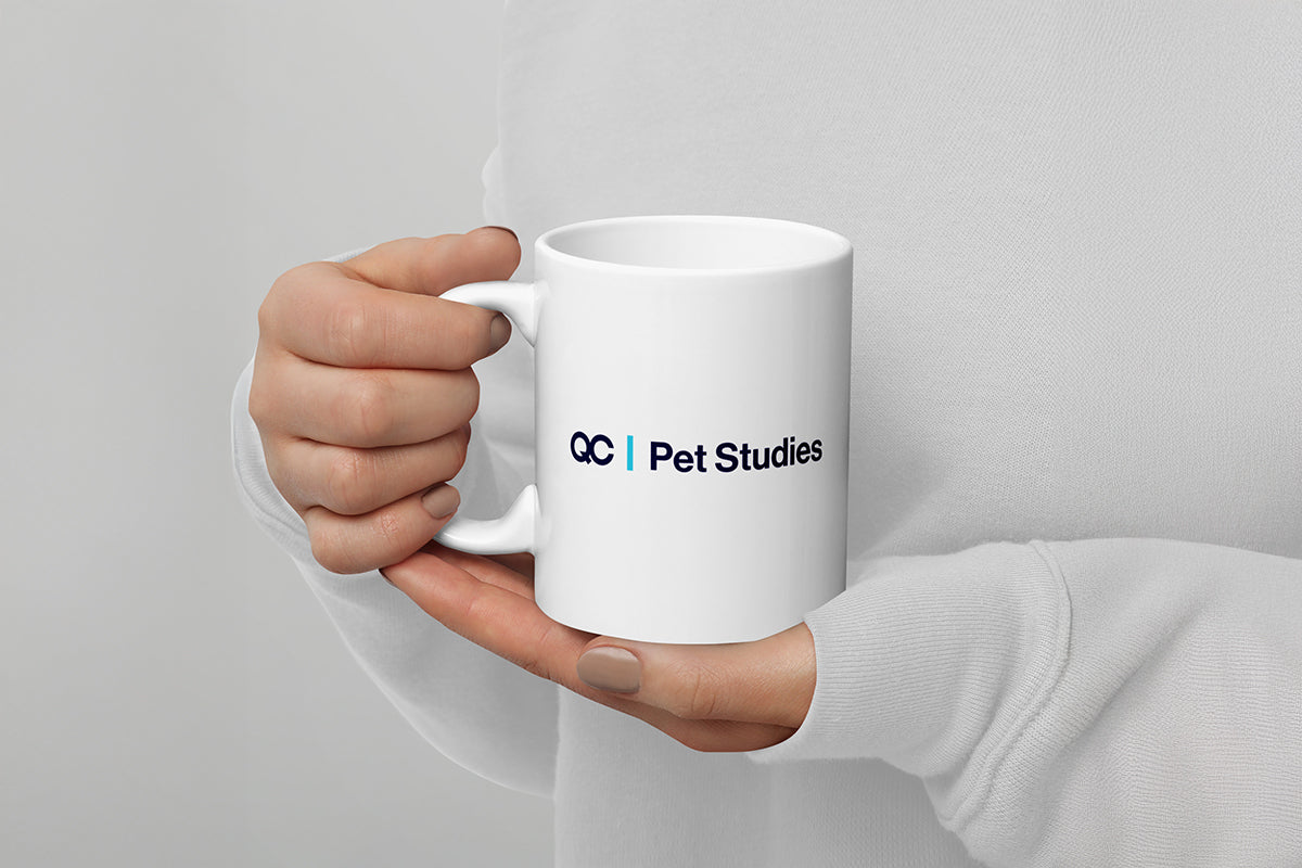 White mug with 'QC Pet Studies' logo held by a person against a gray background