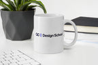 White mug with 'QC Design School' logo on a desk with a plant and books in the background