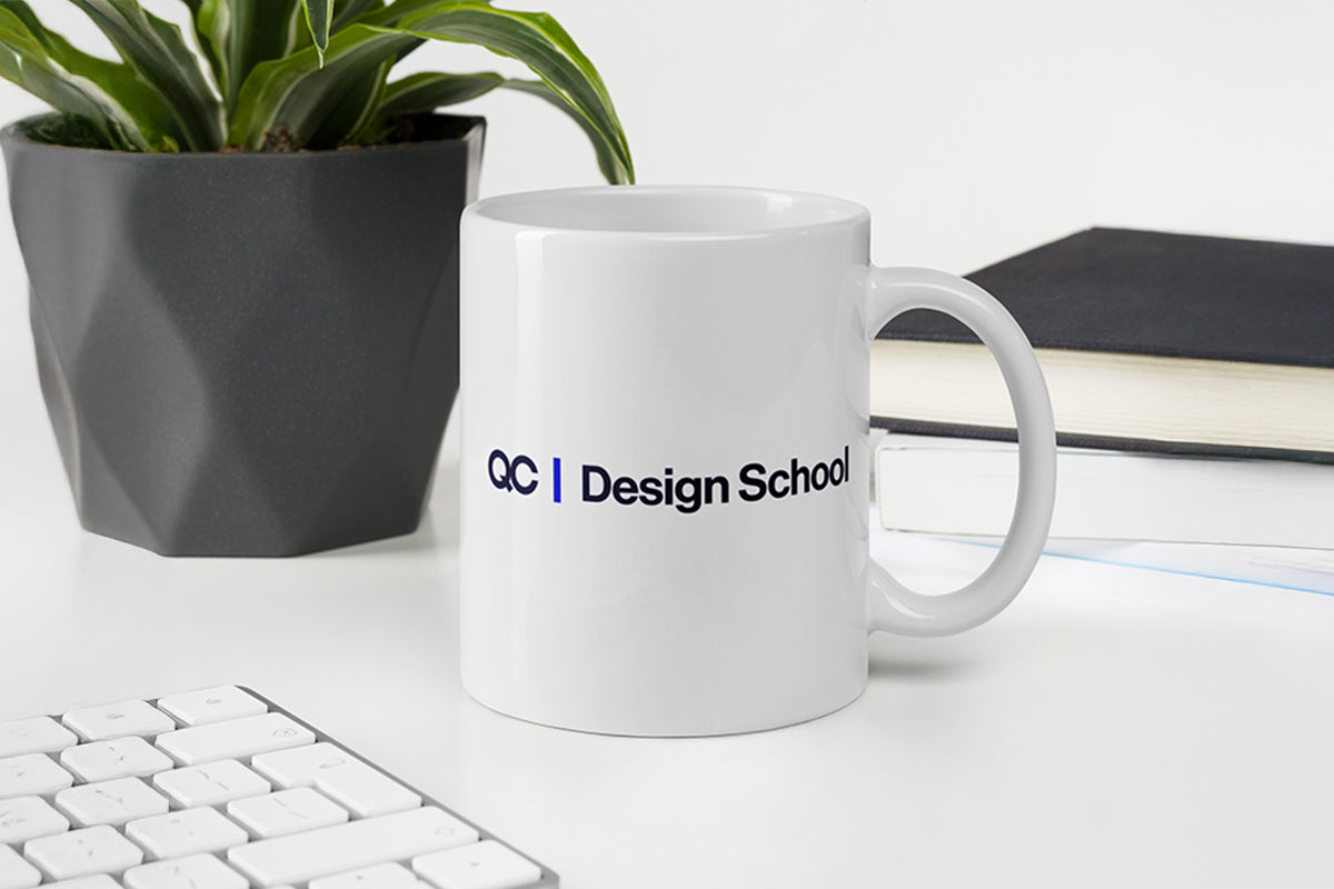 White mug with 'QC Design School' logo on a desk with a plant and books in the background