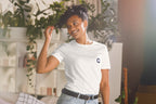 Woman wearing a white t-shirt with a QC logo, standing in a room with plants.