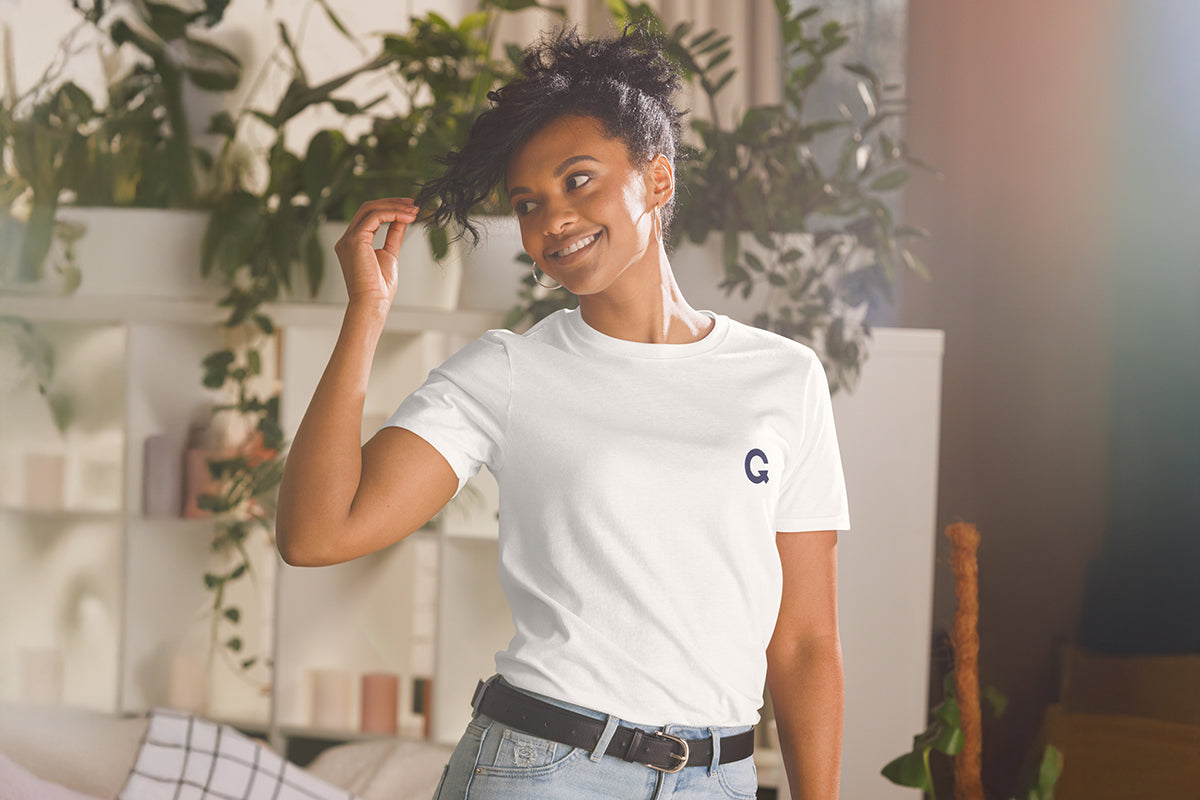 Woman wearing a white t-shirt with a QC logo, standing in a room with plants.