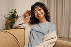 Woman wearing a gray t-shirt with 'QC Design School' text, sitting on a couch.