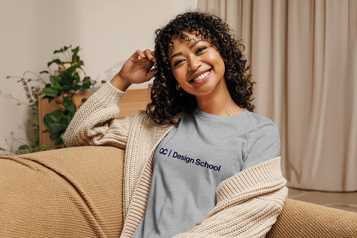 Woman wearing a gray t-shirt with 'QC Design School' text, sitting on a couch.