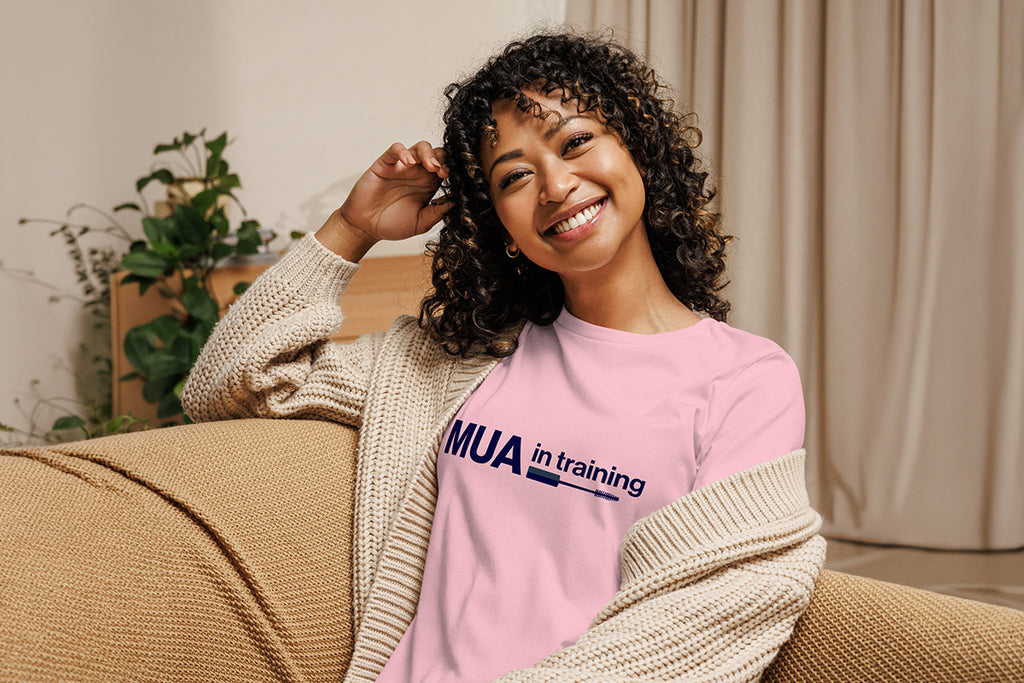 Woman wearing a pink 'MUA in Training' shirt sitting on a couch.