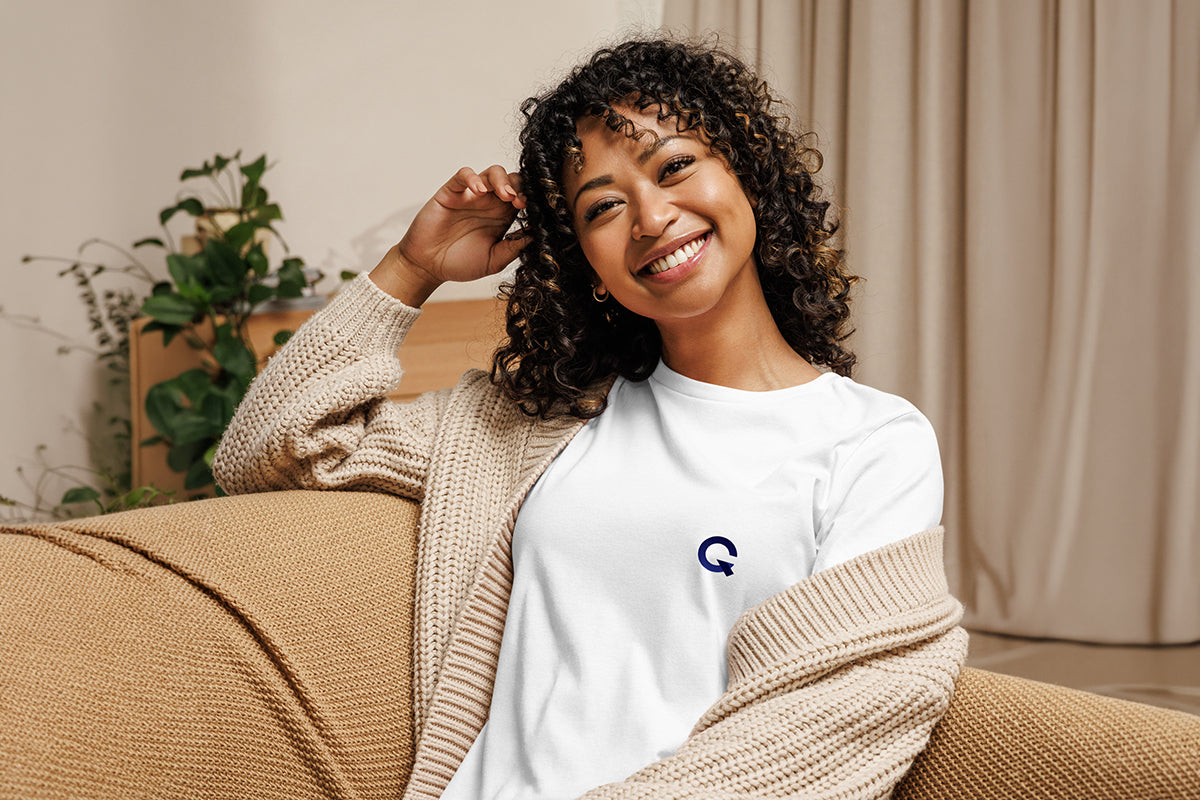 Woman sitting on a couch wearing a white t shirt with a Q logo, smiling.